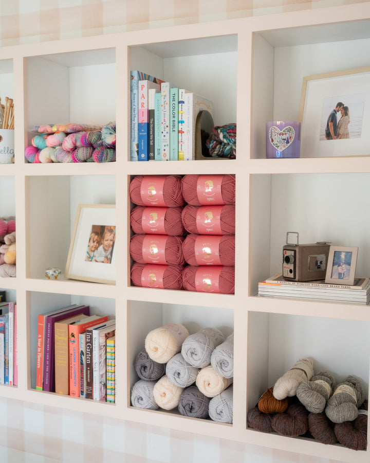 A cozy shelf of rose pink yarn balls nestled among books and sentimental items